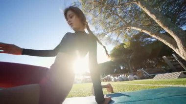 Gün doğumu yogası minderde ısınıyor, sporcu derin nefes alıyor, ağaçlar ve yumuşak güneşli huzurlu bir park, yumuşak hareketlilik