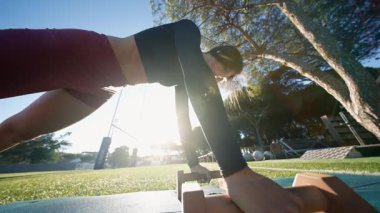 Siyah kadın sporcu, elle desteklenen kalas üzerinde kontrollü bir şekilde dengede duruyor, ağaçların arasından güneş ışığı saçıyor, arka planda park ekipmanları var.,