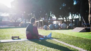 Sunny Park 'ta mindere oturmuş siyah kadın atlet diz bağlarını nazikçe esnetiyor, spor küreleri ve park mimarisi kullanıyor.