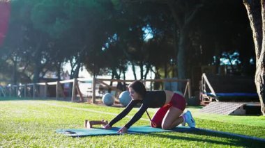 Güneşli parkta paspasa uzanan siyah kadın antrenör, diz çökmüş kedi-inek sekansı, atletik bluz ve şort, egzersiz topları ve ağaçlar.