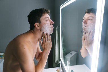 Modern banyodaki LED aynasında tıraş olan üstsüz bir adam, köpük uyguluyor ve çene hattını kontrol ediyor, odaklanmış ifade, cilt bakımıyla batıyor.