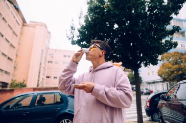 Pembe kapüşonlu genç adam iskan caddesinde sigara içiyor, güneş gözlüğü takıyor ve elinde bir tüp tutuyor, park halindeki arabalarla işe gidiş gelişler gibi.,