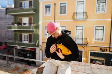 Samimi çatı anı süveter giymiş küçük köpeğe sarılma, odaklanmış bakış, yumuşak güneş ışığı, pastel bina arkaplanı, günlük kıyafet.