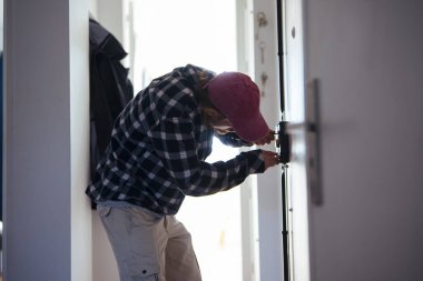 Yerli çilingir iç kapının sürgüsünü ayarlıyor, tornavida ve pense, flanel ceket ve kep, metodik