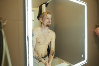 Üstsüz adam aydınlatma aynasında profil kontrol, görünüm ayarlama ve dövmeler görünür, modern ile yakın tarih öncesi tımar anı