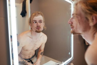 Üstsüz adam aynaya son bir kez bakıyor, deriyi ve dövmeleri kasıtlı hareketlerle inceliyor, hazır olduğunu hissediyor.