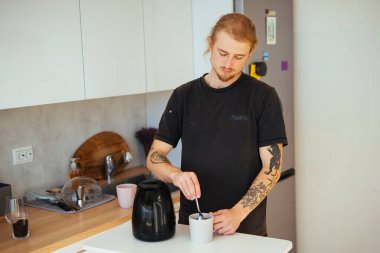 Dövmeli adam, seramik kupanın içinde metal kaşıkla kahve karıştırıyor. Yakınlarda elektrikli su ısıtıcısı, odaklanmış ifade, görünür kol dövmeleri.,