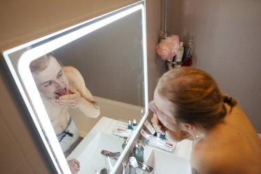 Aydınlatılmış makyaj aynası kullanan, pamuklu ped ile temizleyici kullanan, sakal ve deriyi denetleyen, tuvalet malzemesi kaplı bir adam.