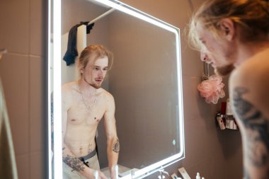 Aynaya bakan üstsüz bir adam, açık sözlü duygusal bir an, kendisiyle yüzleşme ve kabullenme, dövmeli kollar