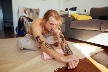 Beyaz adam oturma odasında halıya uzanmış, kolları dövmeli, güneş ışığı ahşap zeminde, sıradan pantolonlar, odaklanmış.