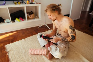 Tişörtsüz dövmeli adam güneşli bir dairede halıda eski bir kamerayı tamir ediyor. Yanında köpük rulo ile yalınayak geziyor, parçalarını söküyor.