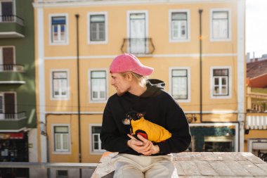 Genç adam köpek yavrusu çatısı çıkıntısına sarılıyor. Renkli apartmanlara bakıyor. Yumuşak öğleden sonra ışığı.