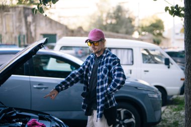 Genç adam açık bagajı işaret ediyor, renkli bavulları konut caddesinde park edilmiş küçük arabaya yüklüyor, flanel gömlek giyiyor.,