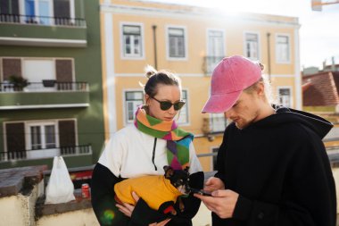 seyahat ikilisi telefonda yönleri kontrol ediyor, güneşli çatı, pastel binalar, gitar kılıfı eğilmiş, süveter giymiş küçük bir köpek, rahatlamış.