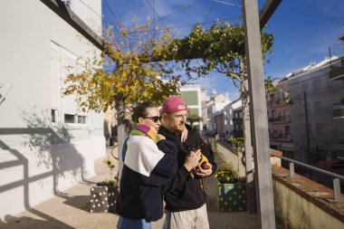Atıştırmalık, renkli kıyafetler ve şehir mimarisi setini paylaşırken güneşli yolda sarılıp gülen iki yakın arkadaş.