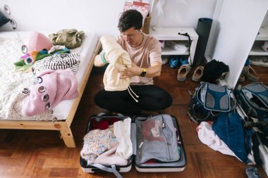 Genç Asyalı adam konferans seyahati için gömlek katlıyor, takım elbise ve kravatları yatağa yerleştiriyor, seyahat programını ve elektronik eşyaları kontrol ediyor.,