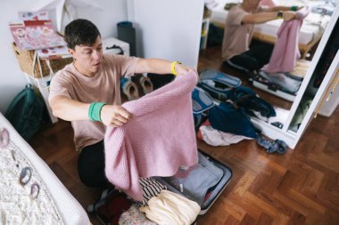 Genç Asyalı adam pembe kazağını dikkatlice topluyor, aile ziyareti için örgü örüyor, hediyesini yumuşak bir çantayla bavula koyuyor.