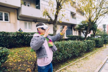 Sanal ses sinyallerini dinlerken VR kulaklığını ayarlayan kişi, hayali kontrollere işaret eden, kentsel balkon arka planı,
