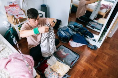 Genç Asyalı öğrenci çalışmak için giysilerini topluyor, çizgili gömleği ve gerekli şeyleri bavula katlıyor, belgeleri düzenliyor.