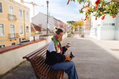 Şehir balkon bankında elinde kahve tutan ve şehre bakan bir kadın, ayaklarda dizüstü bilgisayar çantası, uzaktan çalışma molası, parlak bir öğle arası.