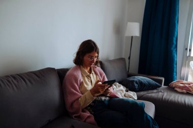 İş molasında telefona bakan bir kadın. Kanepede dinleniyor. Yakınlarda yumuşak bir battaniye, mavi perde arkaplanı.,