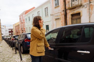Telefonun yanında beyaz bir kadın, elinde anahtarlık, ekranda endişeli bir bakış, kaldırımda pastel boya ile dikiliyor.