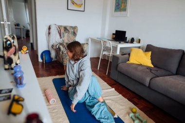 Beyaz bir kız halıya neşeyle atlıyor, paspas ve oyuncaklarla ısınıyor, gri kapüşonlu ve mavi pantolonlu, süper kahraman taklidi yapıyor.