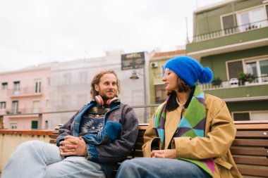 Ahşap bankta oturan çift yansıtıcı sohbetler ve sürekli göz teması kuruyor, şehir balkonuyla yakın çerçeveler kuruyor.