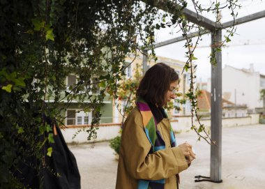 Asılı sarmaşıkların altında duran genç kadın telefon, yan profil ve yumuşak bakışlar derin düşüncelere dalmış, onu takip ediyor.