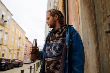 Beyaz adam elinde bira şişesi, desenli süveter, kot ceket, desenli duvar, kaldırım taşı sokak, akşam