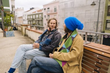 Adam ve yoldaş sırayla sırıtarak oturuyorlar, renkli örgü şapkalar ve paltolar, nehir kenarında pastel binalarla geziniyorlar.,