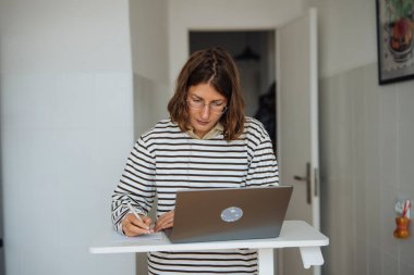 Uzaktan çalışma için dizüstü bilgisayar kullanan, notlar yazan ve odaklanmış duruş ve minimalist evle programı düzenleyen bir kadın.