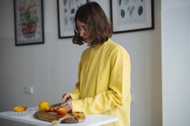 Kadın pencerenin yanında turuncu kesiyor, parlak içerik yaratıyor odaklanmış eller ve yumuşak yan ışık, şık sarı üst