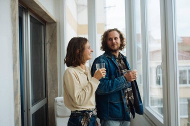 Balkonda şarap içen çift kot ve nötr kıyafetlerle yaratıcı kilometre taşını kutluyor. Gün batımı ışığı, ufuk çizgisi manzarası.,