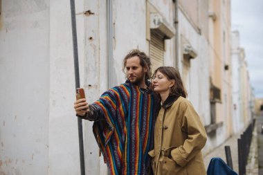 Beyaz çift telefon haritasında yön ve güzergah planlamasını kontrol ediyor. Dar şehir şeritlerinde gezinirken odaklanmış ifadeler var.