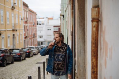 Dar kaldırımlı caddede paket kahve içen adam, tarihi mahallede kot ceketli ve katmanlı sıradan bir gezinti yapıyor.