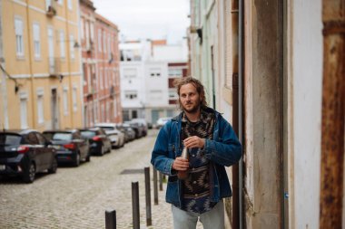 Elinde kamerayla dar bir sokakta duran fotoğrafçı, kot ceket ve eşarp, dikkatli bakış, kaldırımlı şerit ve park halindeki arabalar.,