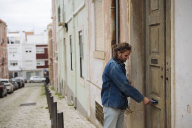 Evin kapısını anahtarla açan adam, yürüme, kot ceket ve sıradan örgü, yıpranmış kapı aralığı.