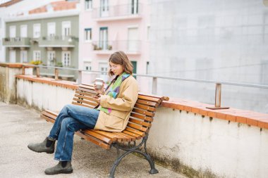 Açık havada kitap okuyan genç bir kadın, kucağında kitap ve defter yığını, odaklanmış duruş ve yumuşak güneş ışığı