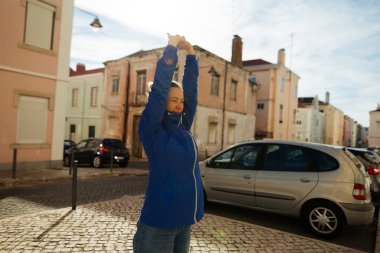 Beyaz adam güneşli meydanda kutlama yapıyor, mavi ceketli kollar havada gökyüzüne gülümserken, kaldırım taşı caddesinde