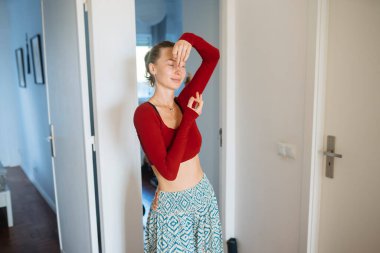 Gülümseyen kadının oyuncu koridor portresi, rahat poz, kapı çerçevesine yaslanma, kırmızı bluz ve desenli etek, samimi yaşam tarzı