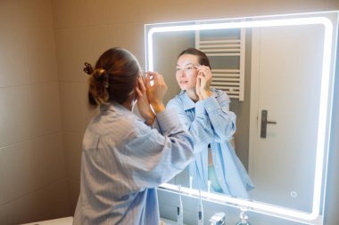 Aynaya göz kremi süren, büyük mavi gömlekli, topuz şeklinde saçı olan, gözlerinin etrafına hafifçe dokunan, LED çerçeveli...