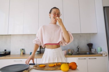 Parlak gülüşü, gamsız enerjisi ve modern mutfaktaki neşeli duruşu olan turuncu dilimi ısıran bir kadın. Pembe eşofman altı eğlenceli bir şeyler ekliyor.