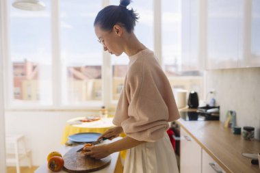 Kafkasyalı bir kadının portakalı doğradığı güneş ışığı tezgahı sahnesi, kesme tahtası, odaklanmış duruş ve hassas hareket, modern mutfak.