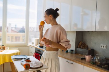 Beyaz bir kadının elinde turuncu ve tablet, uzun bir masada hızlı sağlıklı atıştırmalıklar hazırladığı parlak mutfak anı, güneş ışığı.