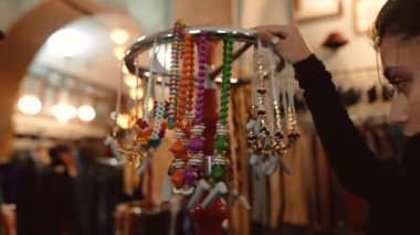 Kafkasyalı kadın butik tezgahında boncuk sergiliyor, satıcı kolye ve tılsım düzenliyor, ellerin kapatılması ve dönüşümlü olarak...