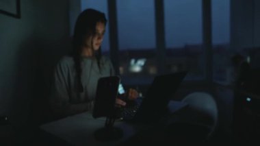 Akşam karanlığında bilgisayarın yanında tablet kullanan kadın çift ekran masa başında, dışarıda yumuşak mavi alacakaranlık, uygulamaları ve mesajları yöneten kadın.,