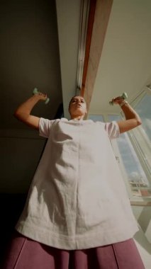 Lowangle kadın açık pencere kenarında küçük ağırlıklar ile esneme, akıcı etek ve güneş ışığı, kollu eğlenceli ev hareketi sekansı