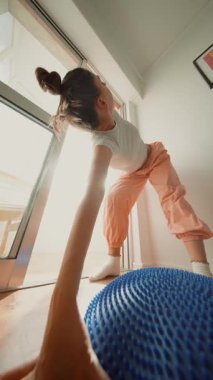 Beyaz kadın, mavi denge yastığına uzanmış koluyla öne doğru eğiliyor. Kendine güvenen kontrollü hareket, güneşli zemin ve geniş.