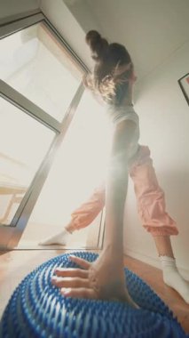 Beyaz kadın, güneş ışığı odasında mavi denge yastığına uzanıp uzanıyor, kollarını uzatıyor ve hareketlerini kontrol ediyor.